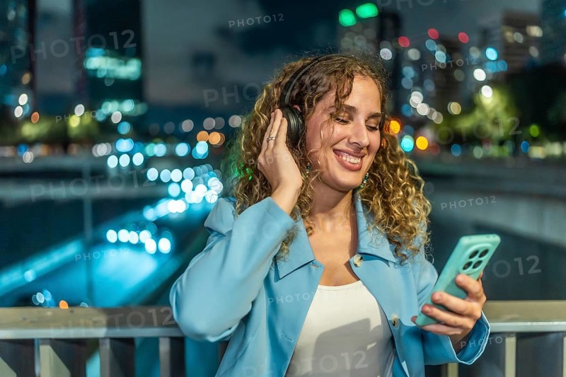 Woman with headphones and smartphone enjoying music while smiling cheerfully, standing on a bridge with city lights blurring in the background at night