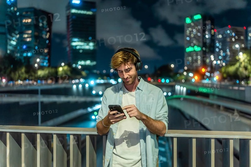 Young man wearing headphones and casual clothing standing on a city bridge at night, smiling and interacting with his smartphone against an urban skyline background