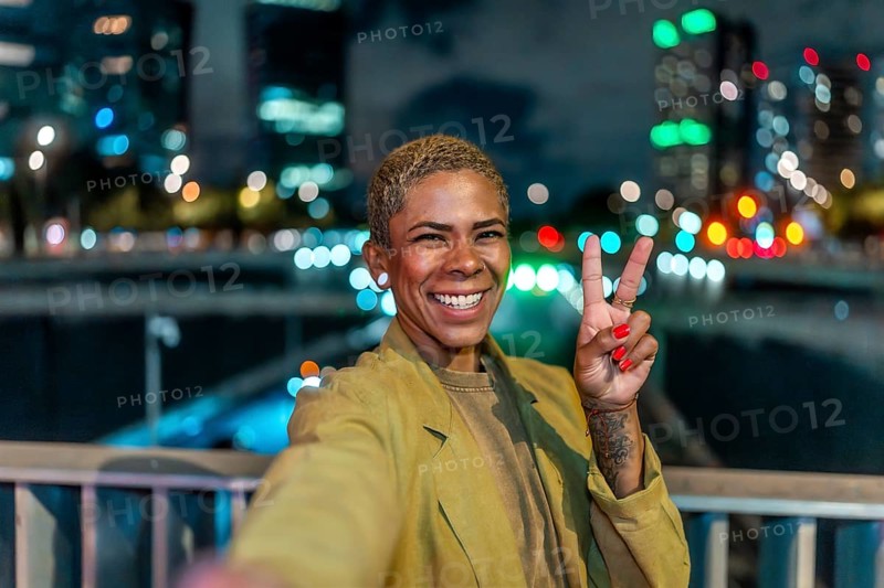 Smiling woman holding up a peace sign, capturing a cheerful selfie moment on a bridge overlooking the illuminated cityscape and vibrant bokeh lights at night