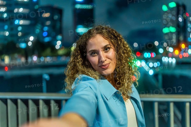 Young woman with curly hair extending her arm to take a selfie, smiling to the camera with blurred city lights and building reflections creating a vibrant bokeh background at night