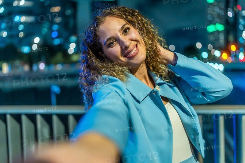 Young woman with curly hair smiling brightly and playfully taking a selfie at night, with a blue jacket and blurred city lights forming a colorful bokeh background