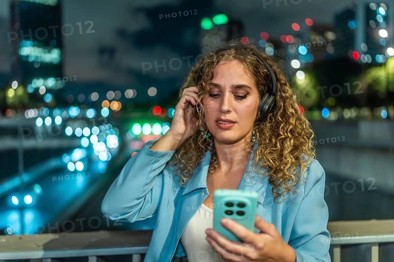 Young woman with curly hair wearing headphones and looking at her smartphone while enjoying music on an urban bridge at night with blurred city lights in the background