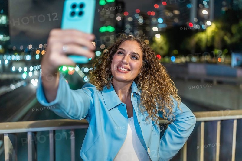 Young woman with curly hair smiling happily while capturing a selfie on her mobile phone, standing on a bridge overlooking city lights and traffic at night