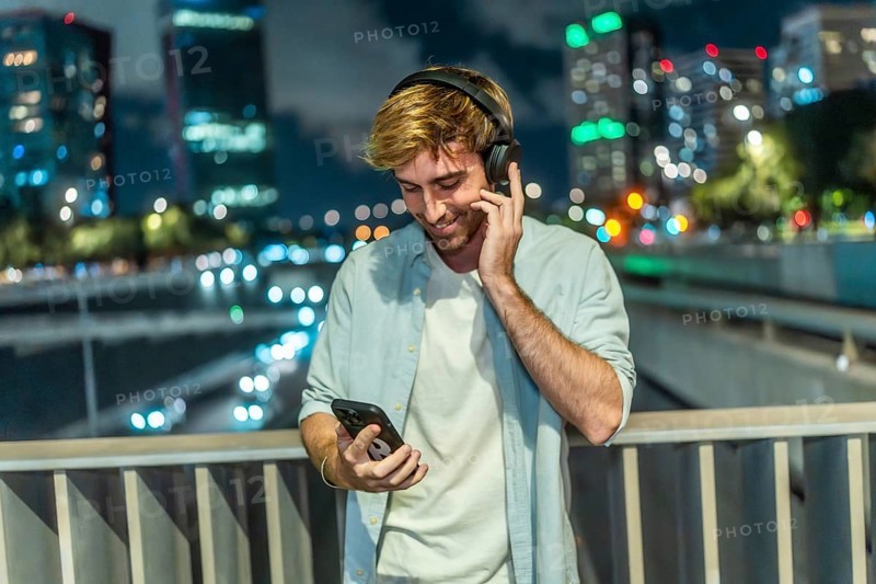 Young man smiling and listening to music through wireless headphones, holding a smartphone while standing on a bridge overlooking a busy city street at night