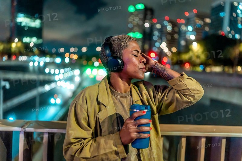 Woman with short hair, wearing headphones and holding a travel mug, relaxing with closed eyes on an urban bridge at night with city lights and bokeh in the background