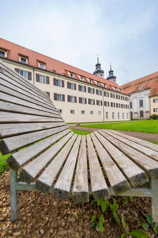 A wooden park bench in the foreground with a monastery building in the background under a cloudy sky, ZFP Zwiefalten, Reutlingen district, Germany