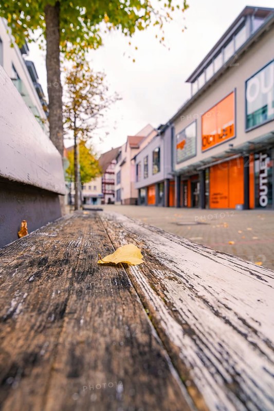 A quiet shopping street with modern buildings, autumn leaves and a wooden bench in the foreground, Nagold, Black Forest, Germany