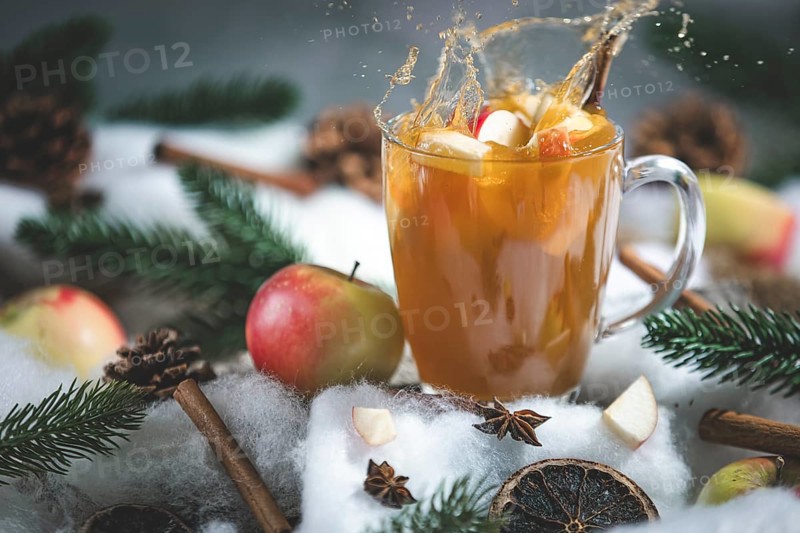 Apple punch splashes out of a glass cup with apple pieces, surrounded by cinnamon sticks and pine branches in a wintry environment