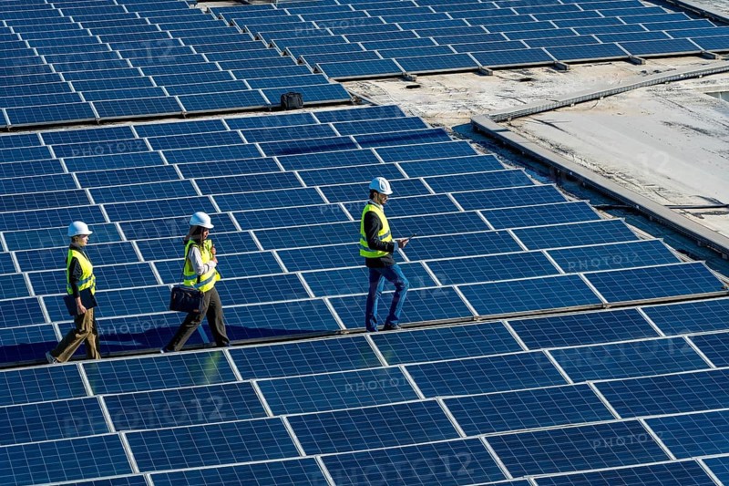 Engineers wearing safety helmets and vests are walking across a large array of solar panels, conducting an inspection of the renewable energy installation on a rooftop