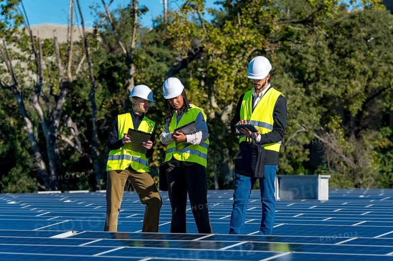 Three diverse engineers wearing hard hats and high visibility vests are inspecting solar panels on a rooftop, using a tablet and clipboard, contributing to renewable energy sustainability