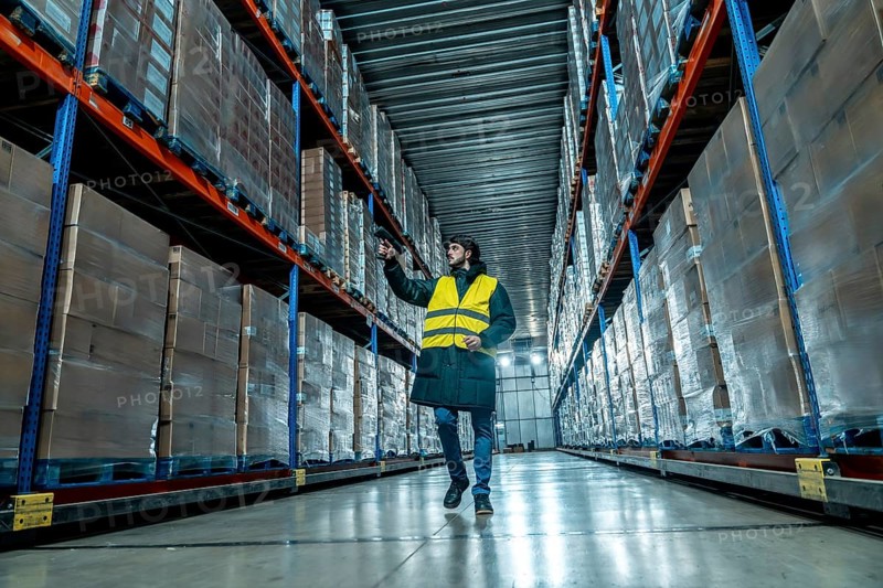 Worker in protective clothing and safety vest holding a barcode scanner, checking products in a temperature controlled logistics facility with tall shelves filled with wrapped pallets