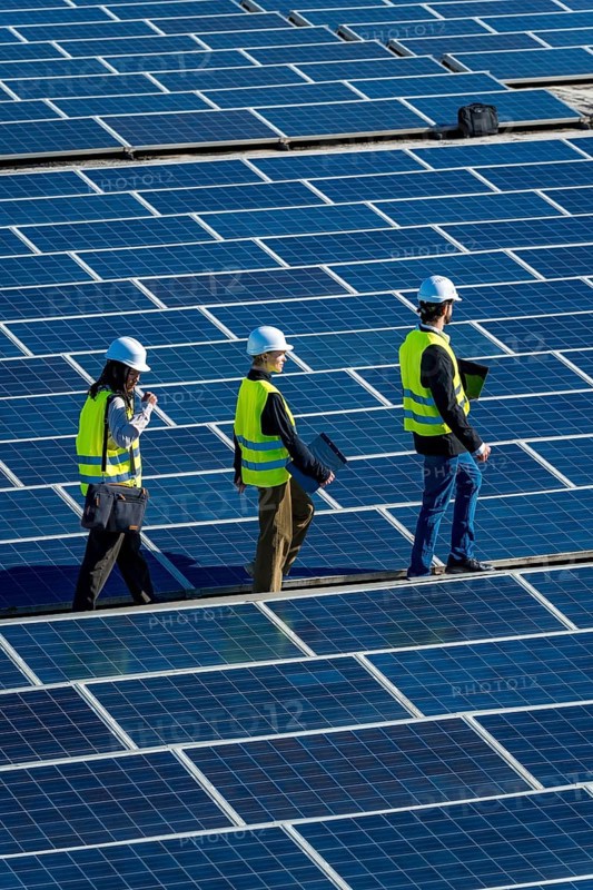 Engineers wearing safety vests and hard hats are walking across a vast array of solar panels at a renewable energy power station, conducting inspection and maintenance work