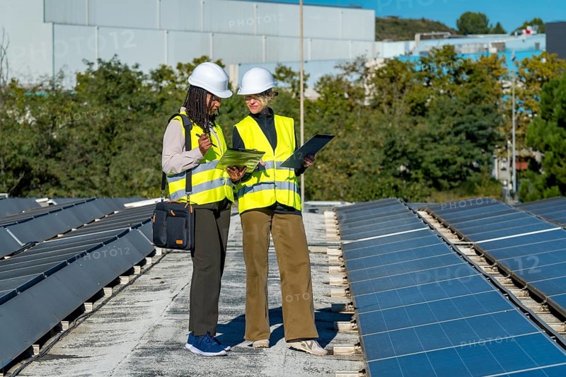 Two female engineers wearing hard hats and reflective vests are reviewing documents while standing on a rooftop next to solar panels, highlighting renewable energy and sustainability
