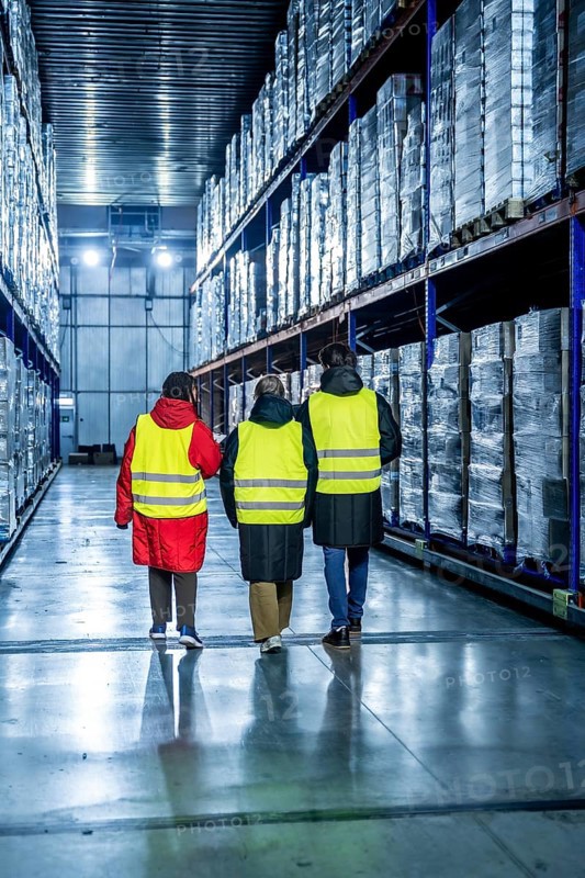 Workers wearing safety vests walking down an aisle in a cold storage warehouse, inspecting pallets of products wrapped in plastic on high racks, managing the supply chain