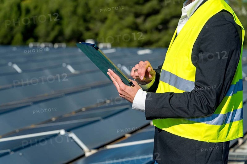 Engineer in safety vest and suit jacket holds clipboard and pen while inspecting and recording data from a large outdoor solar panel array on a sunny day, assessing performance and maintenance
