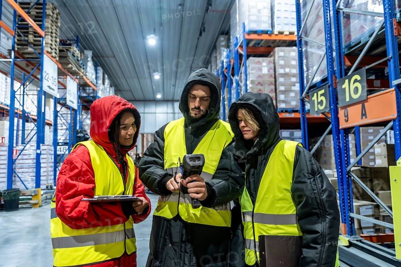 Three warehouse workers in protective clothing and high visibility vests are collaborating while scanning products in a cold storage facility, managing inventory and supply chain operations