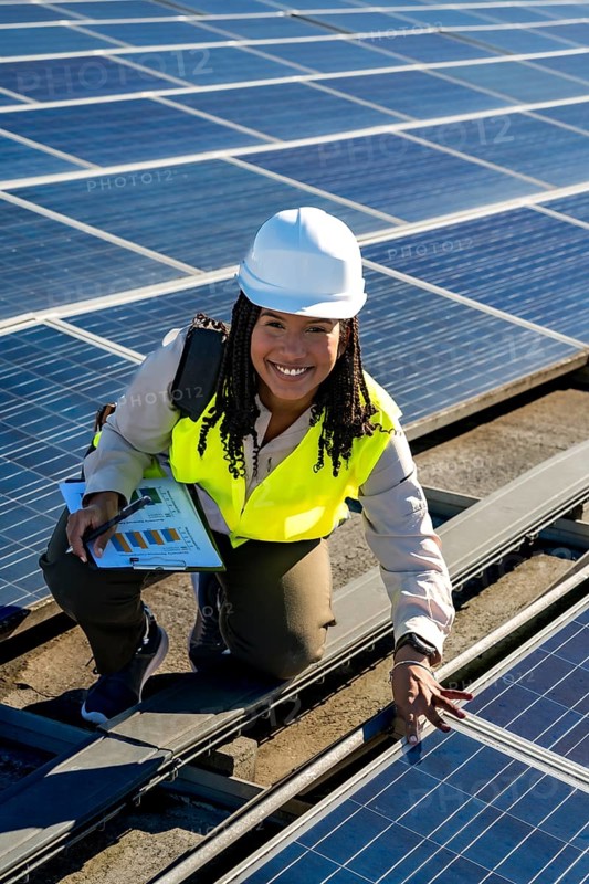 Young woman engineer with safety helmet and vest, smiling while inspecting photovoltaic panels installation on a sunny rooftop, working on sustainable energy and green power
