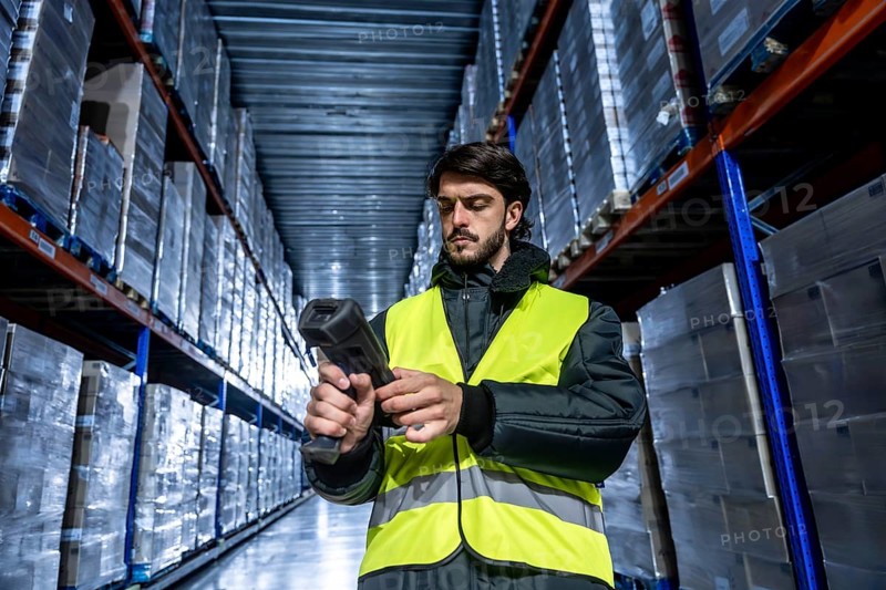 Worker wearing a safety vest and jacket operating a barcode scanner, managing inventory and shipping logistics inside a large industrial cold storage warehouse with tall shelves and packed goods
