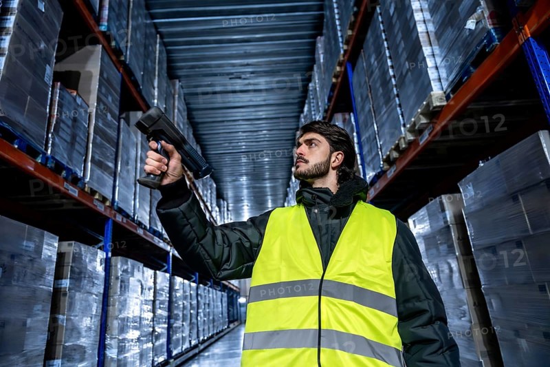 Worker in a safety vest and warm jacket performing inventory management, scanning product barcodes on high shelves in a large cold storage or frozen logistics warehouse