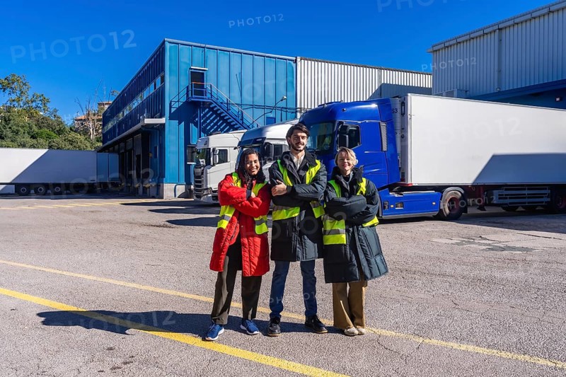 Logistics team members smiling, wearing safety vests and warm clothing, standing outdoors with arms crossed at a frozen storage warehouse with parked refrigerated trucks