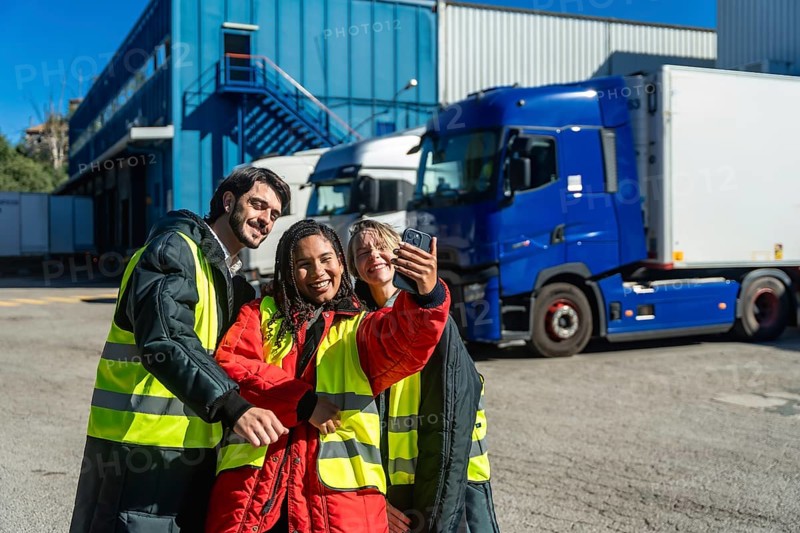 Diverse logistics team members smiling and taking a selfie outside a cold storage warehouse with trucks, symbolizing teamwork and employee satisfaction in transport and supply chain