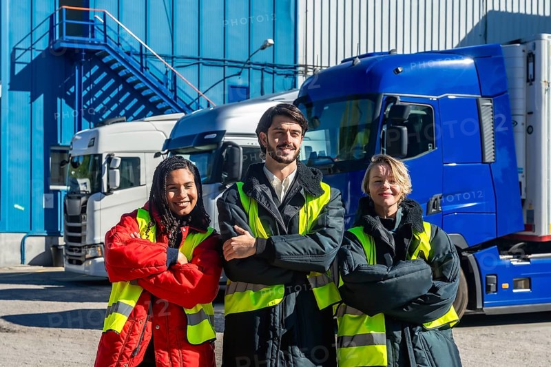 Diverse logistics team in high visibility vests and insulated jackets standing with arms crossed outside a cold storage facility, with refrigerated trucks behind, proud and professional