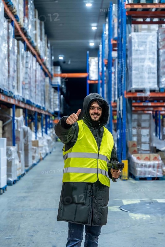 Warehouse worker in warm clothing and safety vest holding a barcode scanner and giving thumbs up inside a cold storage facility, smiling during inventory and logistics operations