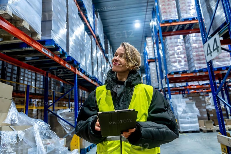 Woman worker managing cold storage logistics, wearing a warm jacket and reflective vest, holding a digital tablet while checking inventory in a large warehouse filled with stacked pallets