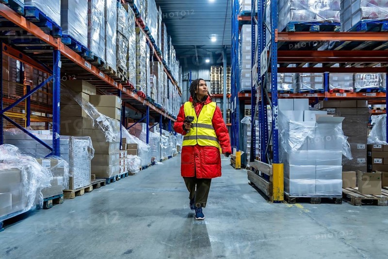 Female warehouse worker wearing warm clothing and a safety vest, scanning inventory while walking through a large industrial cold storage facility filled with pallets of wrapped goods on high racks