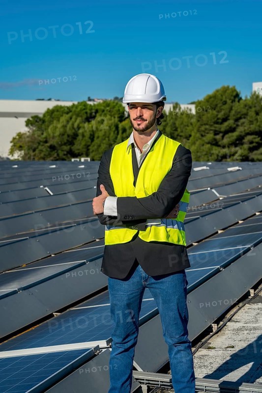 Engineer wearing a hard hat and safety vest standing confidently with arms crossed on a rooftop solar panel array, representing sustainability and green energy development