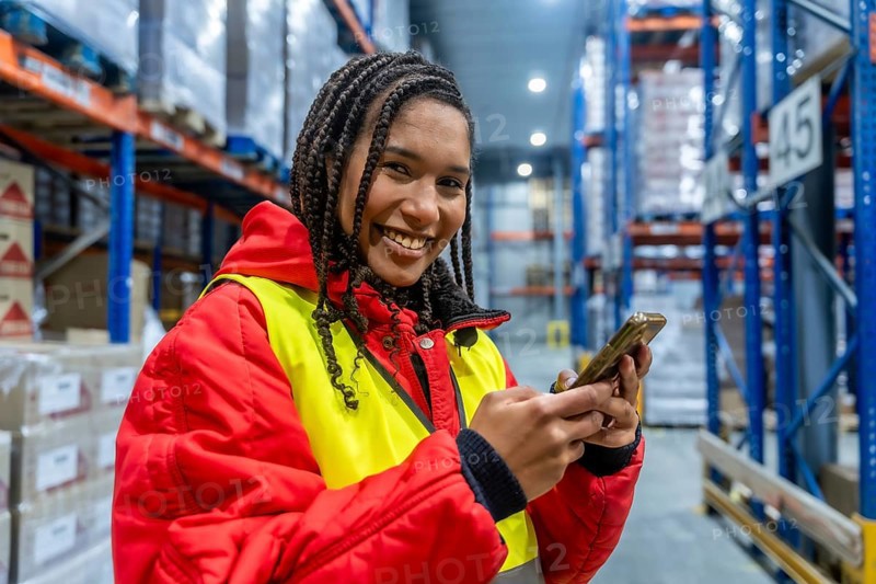 Smiling female worker managing cold storage inventory, checking orders and ensuring efficient logistics operations for frozen goods using a mobile device in a large industrial warehouse