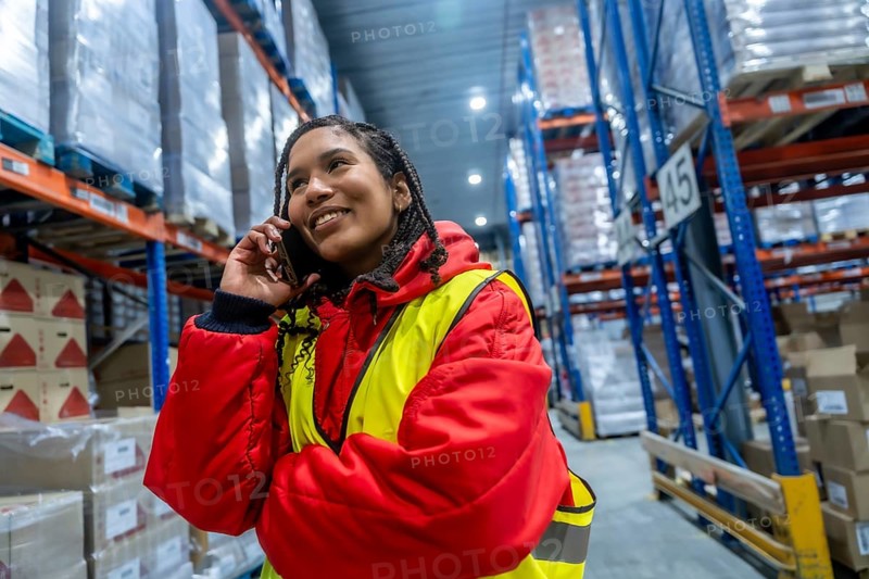 Woman worker smiling while talking on a mobile phone, wearing warm workwear in a vast cold storage warehouse, managing supply chain and frozen food logistics