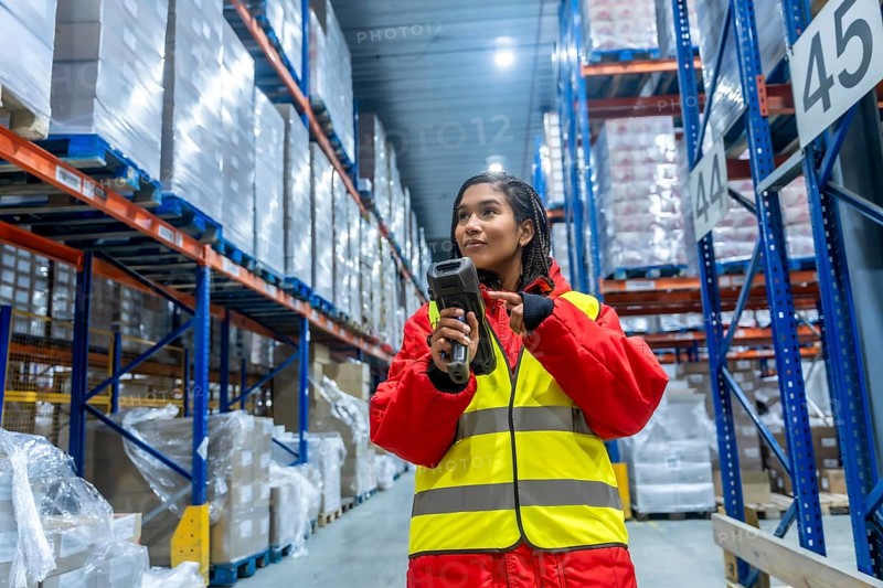 Woman wearing protective cold weather gear and a high visibility vest operating a barcode scanner, managing inventory and logistics in a large, modern cold storage facility