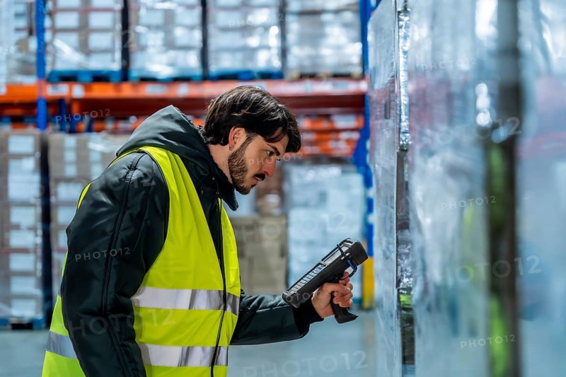 Worker wearing protective clothing and high visibility vest operating a barcode scanner to check inventory on pallets in a frozen distribution warehouse, managing cold chain logistics