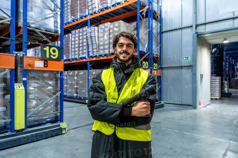 Man in safety vest and warm clothing holding a scanner, working in a cold storage warehouse with tall racks of frozen goods for logistics and supply chain operations