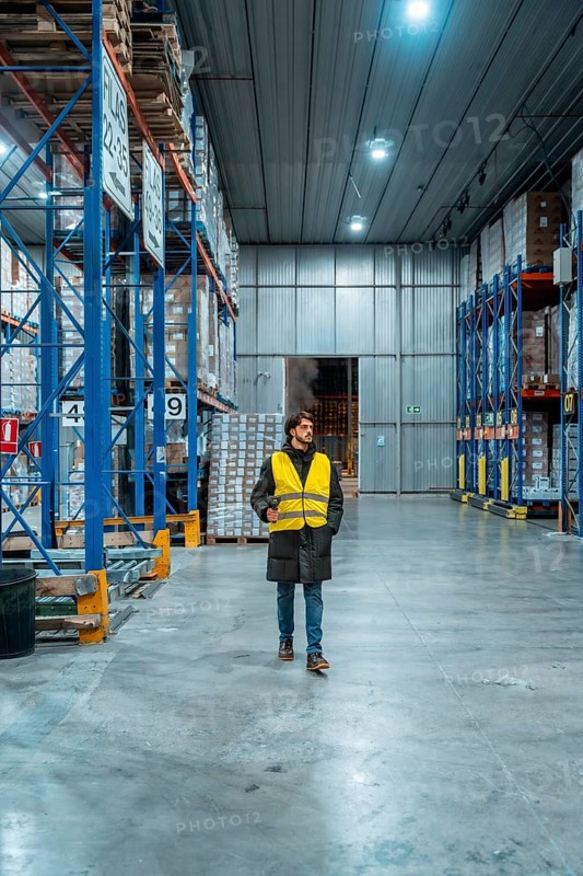Male worker wearing a safety vest and winter jacket managing inventory in a large, modern cold storage warehouse, ensuring efficient frozen logistics and supply chain operations