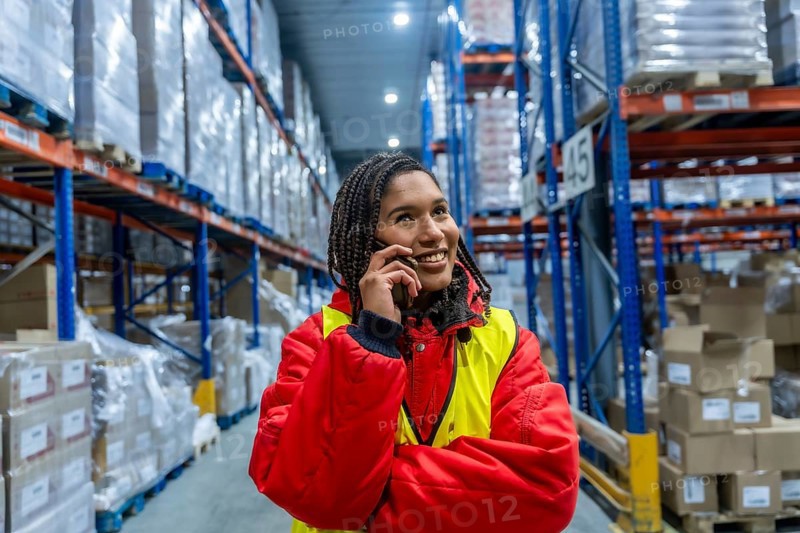 Woman warehouse worker wearing warm clothing and a safety vest, smiling while having a phone conversation inside a large cold storage facility, managing logistics and inventory