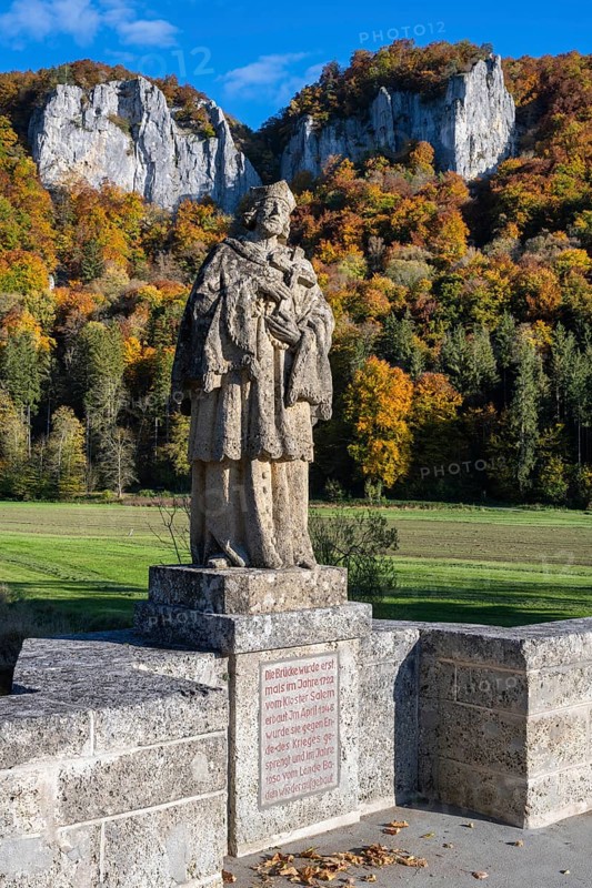 Statue, saint statue of Saint Nepomuk, patron saint of bridges, bridge saint on the historic arched bridge, Hausener Brücke behind the distinctive Hausener Zinnen, climbing rocks, Jurassic limestone rocks, Weissjura, Jurassic rocks, limestone rocks, surrounded by autumnal vegetation, Hausen im Tal, Upper Danube Valley, Baden-Württemberg, Germany