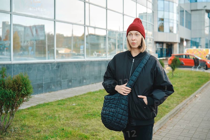 A woman wearing a black jacket and red beanie walks along a modern glass building. She holds a large bag and seems to enjoy a day in the city, surrounded by greenery and urban scenery