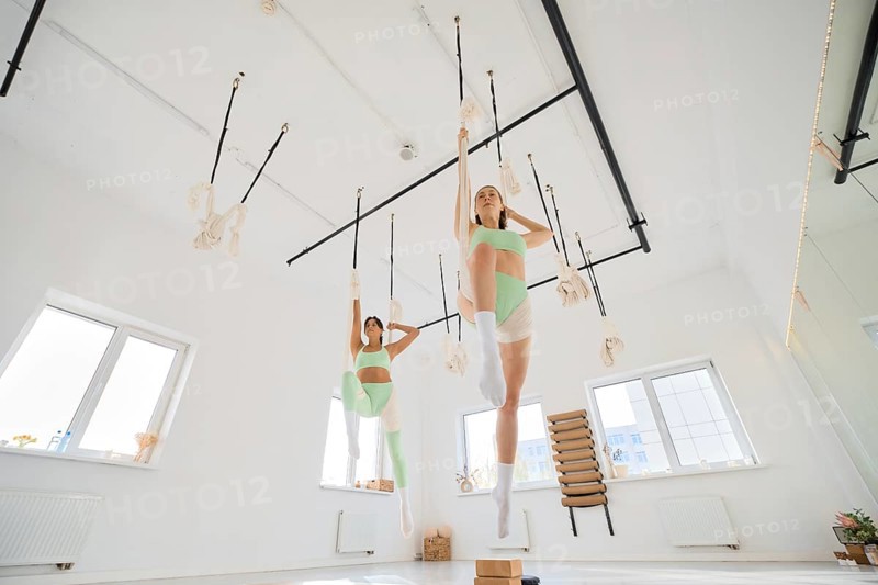 Two individuals engage in a fitness stretching routine using a hammock in a bright, spacious studio. The atmosphere is energetic as they focus on flexibility and strength training