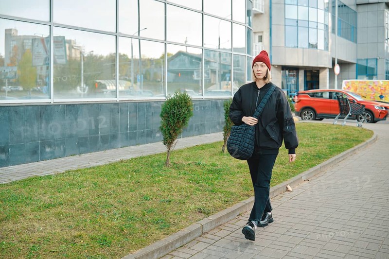 A woman strolls along a wide walkway surrounded by a mix of urban buildings and greenery. She wears a black jacket, black pants, and a red beanie while carrying a shoulder bag