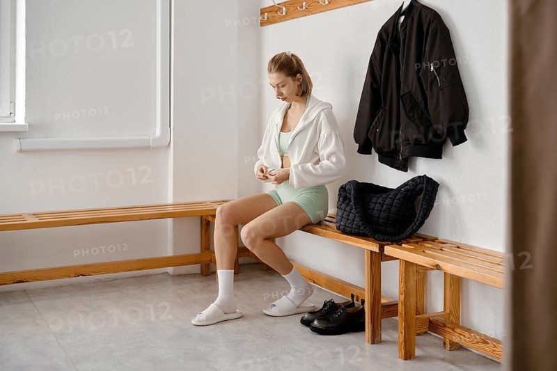 A young woman in a light green outfit sits on a wooden bench in a gym locker room. She is putting on her hoodie while a black jacket hangs nearby. A gym bag rests on the bench beside her