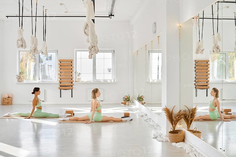 In a spacious yoga studio filled with natural light, two women perform stretches on mats. The room features large mirrors, calm decor, and hanging ropes
