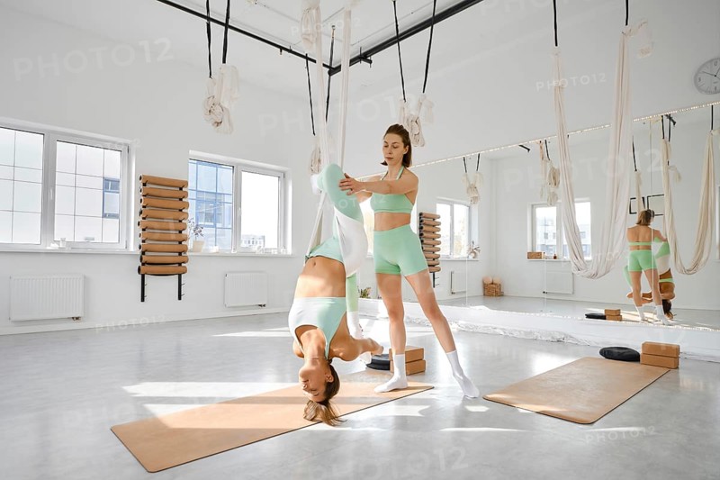 Two fitness enthusiasts are participating in a stretching session using a hammock in a bright, airy studio