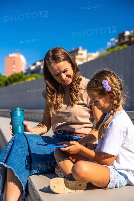 Mother and daughter smiling, sitting together on steps enjoying time outdoors, holding a reusable cup and interacting with a digital tablet, learning and connecting in sunlight