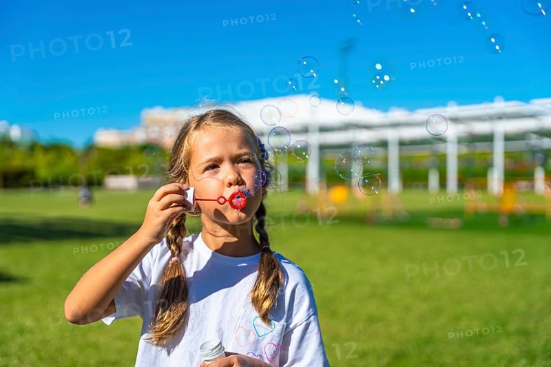 Child with pigtails enjoying summer playtime outdoors, actively creating and watching soap bubbles float in the bright blue sky above a green grass field