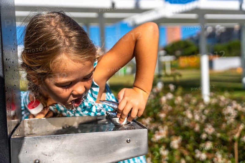 Little girl bending to drink cool water from a stainless steel park fountain on a sunny summer day, enjoying refreshment, hydration and playful outdoor fun amidst greenery