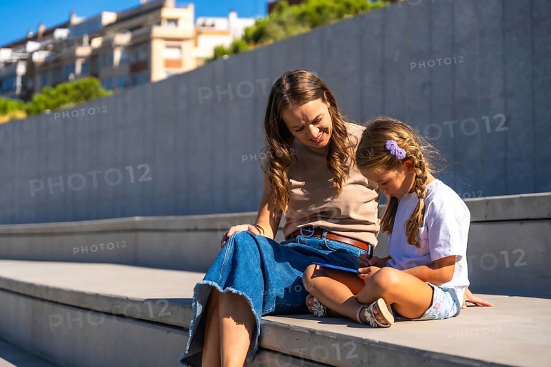 Mother and daughter sitting on concrete steps, sharing a tablet and engaging in an educational activity, enjoying their time together outdoors on a sunny day