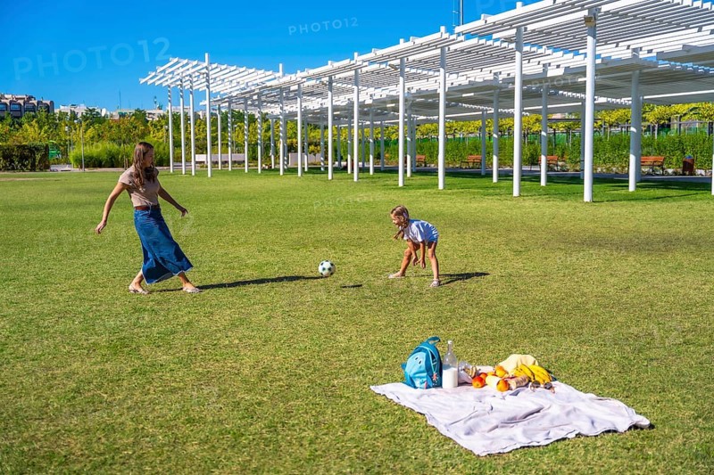 Mother and daughter playing soccer in a sunny green park, enjoying a picnic with blanket and snacks, bonding, active recreation and summer family fun outdoors