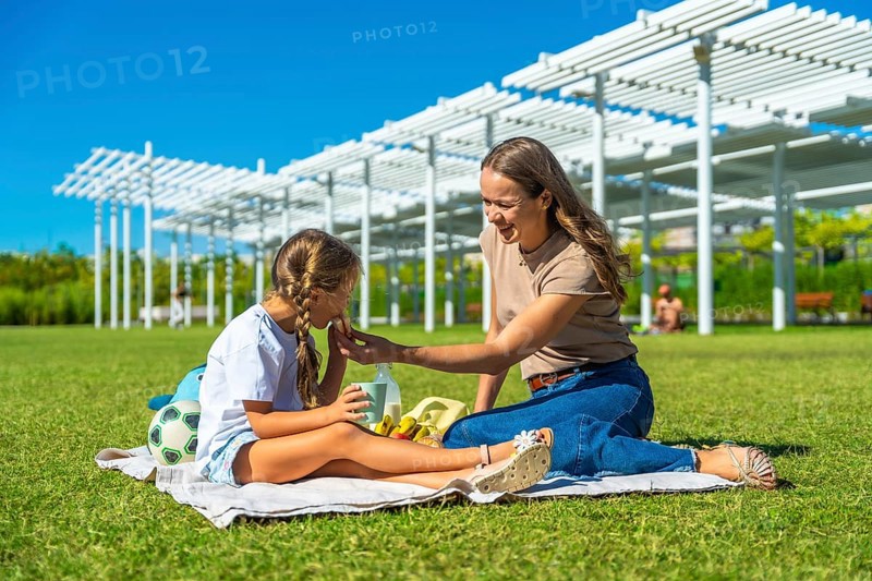 Mother and daughter enjoying a sunny summer picnic on green grass, sharing food and snacks while relaxing outdoors under a blue sky, fostering family bonding
